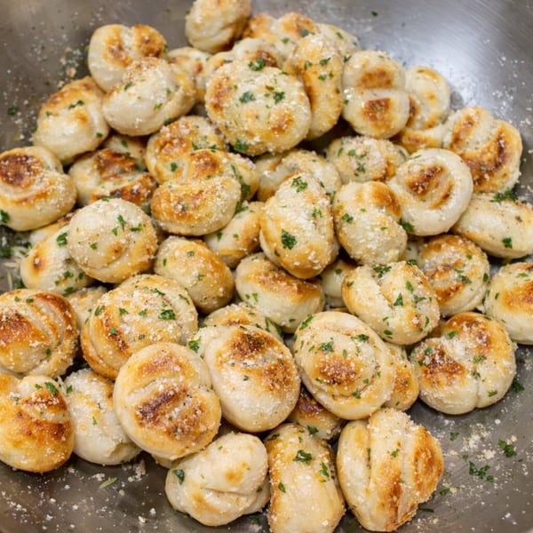 A large metal bowl filled with golden-brown garlic knots, sprinkled with parmesan cheese and parsley.